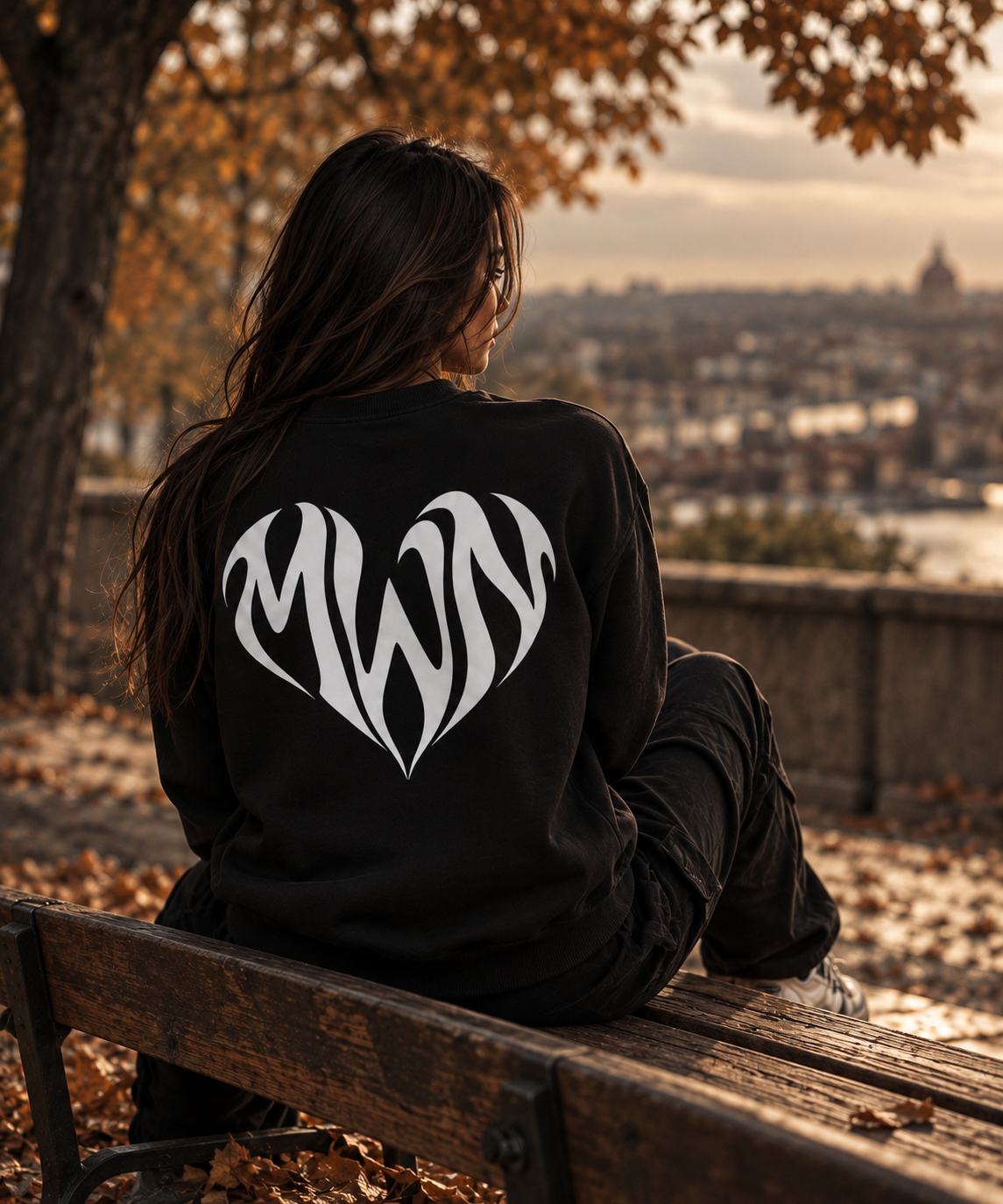 Sweat noir MWN HEART porté de dos par une femme assise sur un banc, grand logo cœur blanc.
